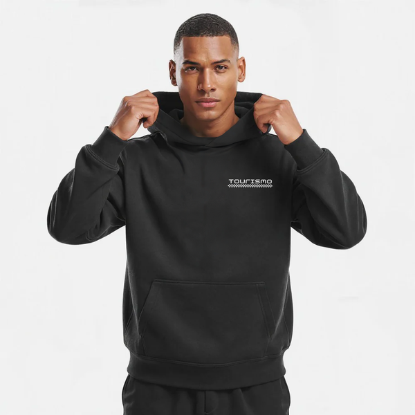 Sweat à capuche noir sans cordon pour homme, modèle "Pression Maximale" signé Tourismo Clothing. Hoodie coupe droite avec poche kangourou, capuche structurée tenue fermement par les deux mains du mannequin. Logo brodé blanc "TOURISMO" avec damier graphique sur la poitrine gauche. Fond studio blanc, cadrage buste, regard frontal intense. Vêtement streetwear inspiré de l’univers automobile, entre technicité textile et attitude déterminée. Idéal pour un look racing discret et puissant.